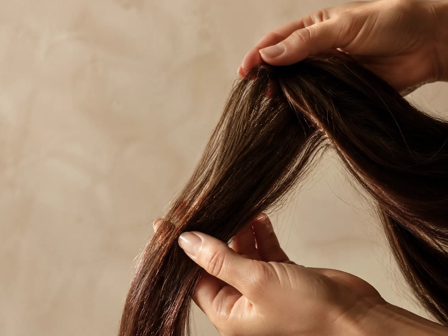 A stylist's hands lifting a length of glossy dark hair against a warm cream plaster background