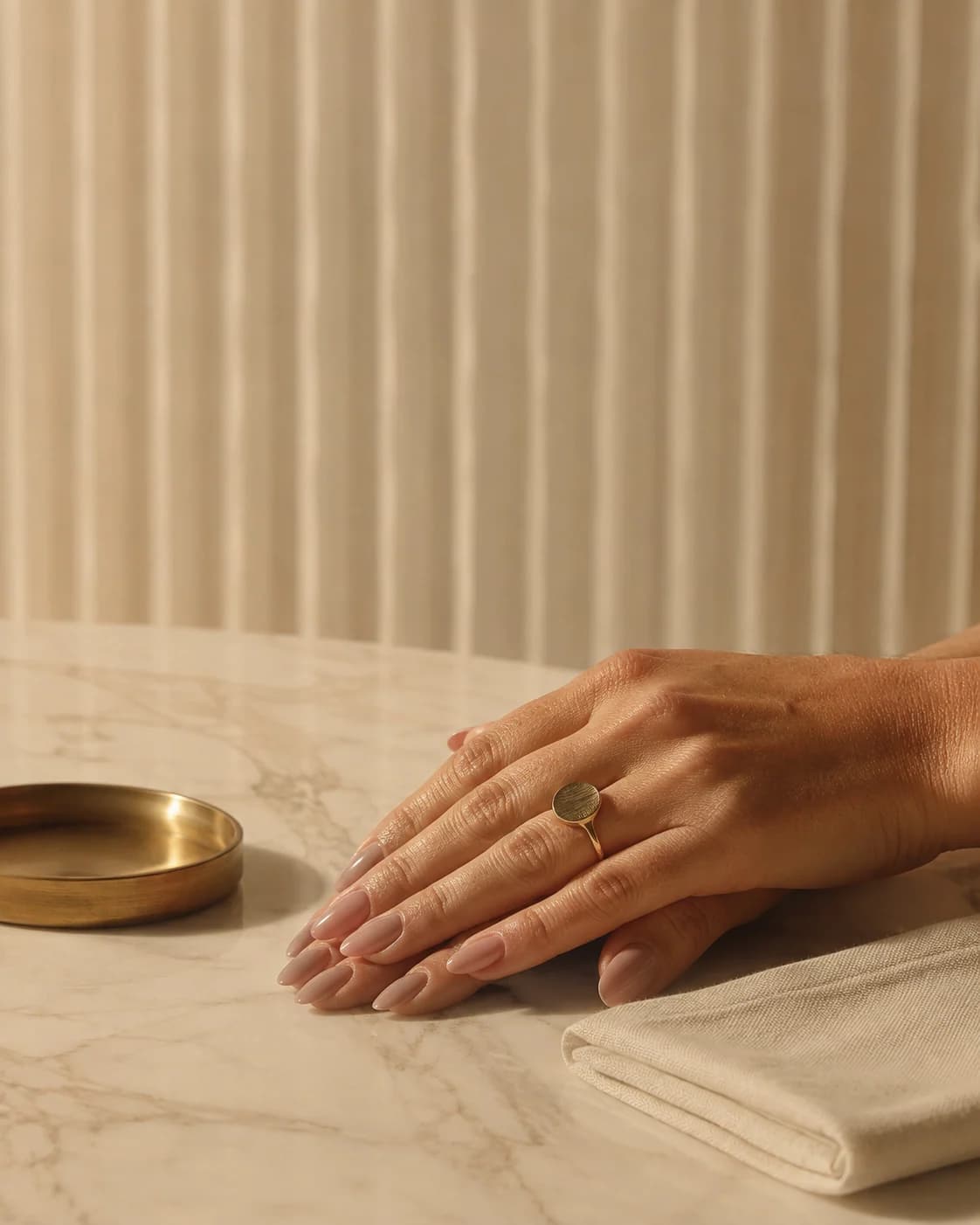 A woman's hands resting on pale marble, nails finished in a soft neutral shade, beside a brushed-brass dish and folded linen